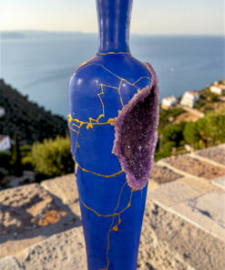Cobalt blue vase with gold crackle glaze and a purple geode cluster, set on a rocky overlook by the sea.