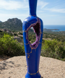 Tall blue ceramic vase sculpture with gold crack lines and a purple crystal ring in its midsection, set on a gravel pedestal with hills and sea in the background.