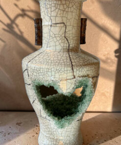 Cream crackled ceramic vase with two dark brown handles and a large broken opening near the base revealing turquoise-green crystalline glaze inside, on a dusty surface.