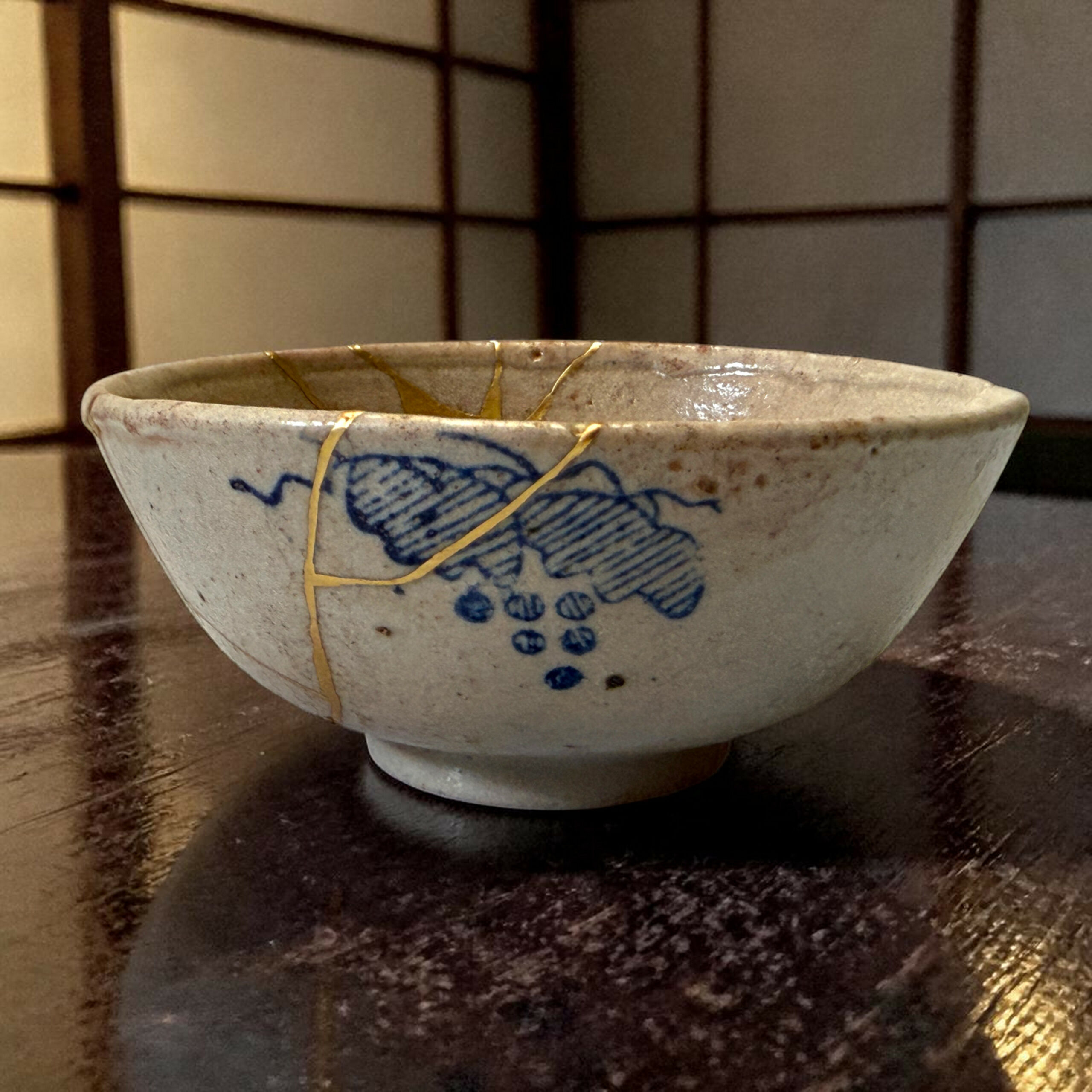 IMG_1573-Photoroom | Kintsugi by Myriam GREFF Close-up of a white ceramic bowl with blue underglaze design and a yellow string draped over its rim on a dark wooden table surface.