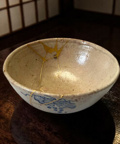 Close-up of a ceramic bowl with blue glaze patterns and gold kintsugi repair seams on a dark wooden table.
