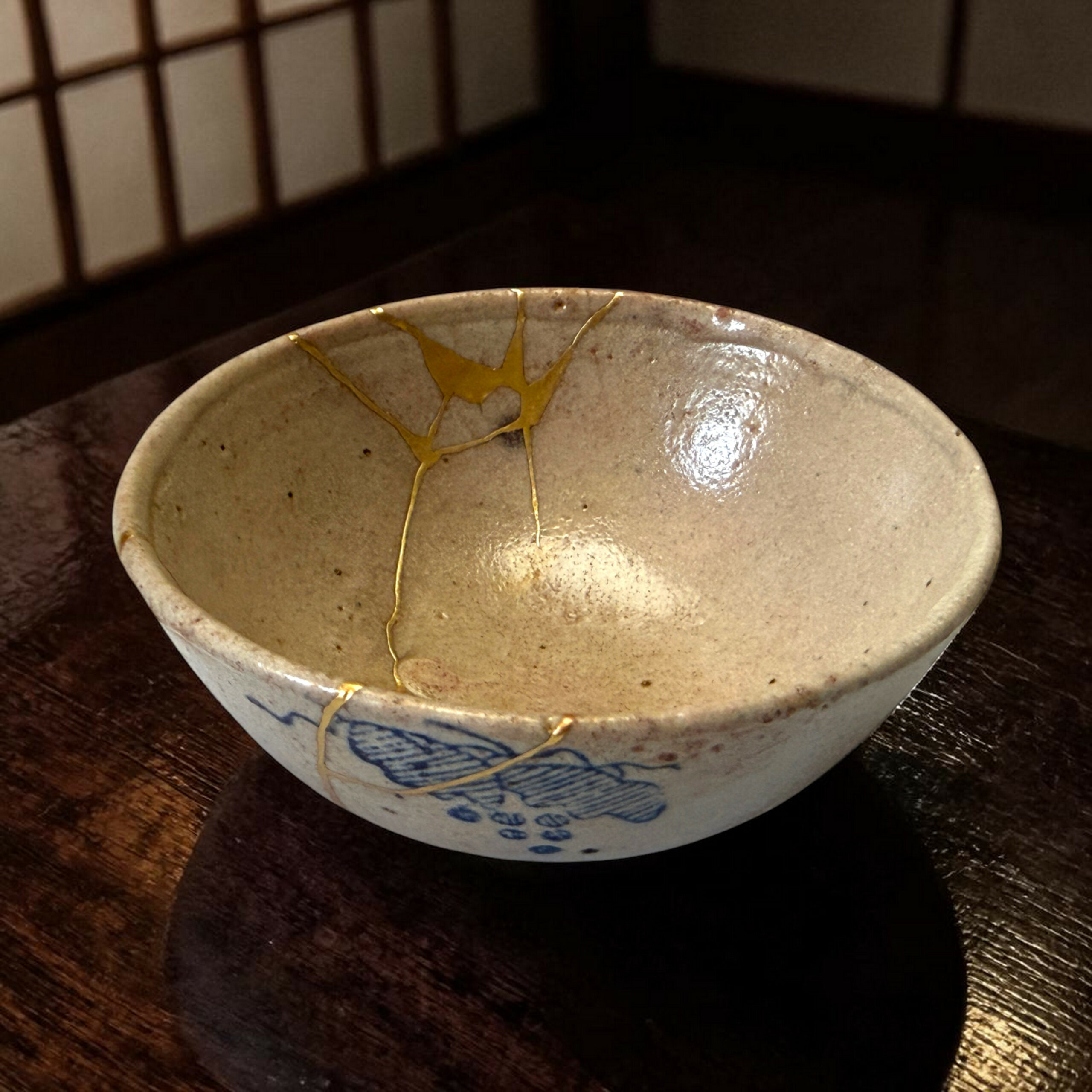 IMG_1574-Photoroom | Kintsugi by Myriam GREFF Close-up of a ceramic bowl with blue glaze patterns and gold kintsugi repair seams on a dark wooden table.
