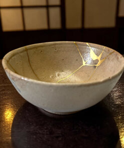 Ceramic bowl with gold-kintsugi cracks on a dark wooden table under warm lighting.
