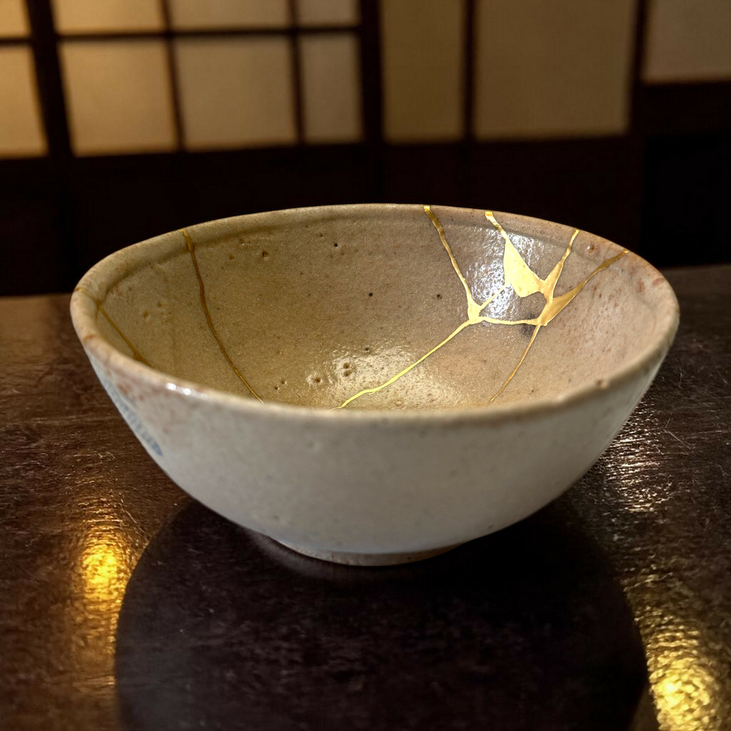 IMG_1575-Photoroom | Kintsugi by Myriam GREFF Ceramic bowl with gold-kintsugi cracks on a dark wooden table under warm lighting.