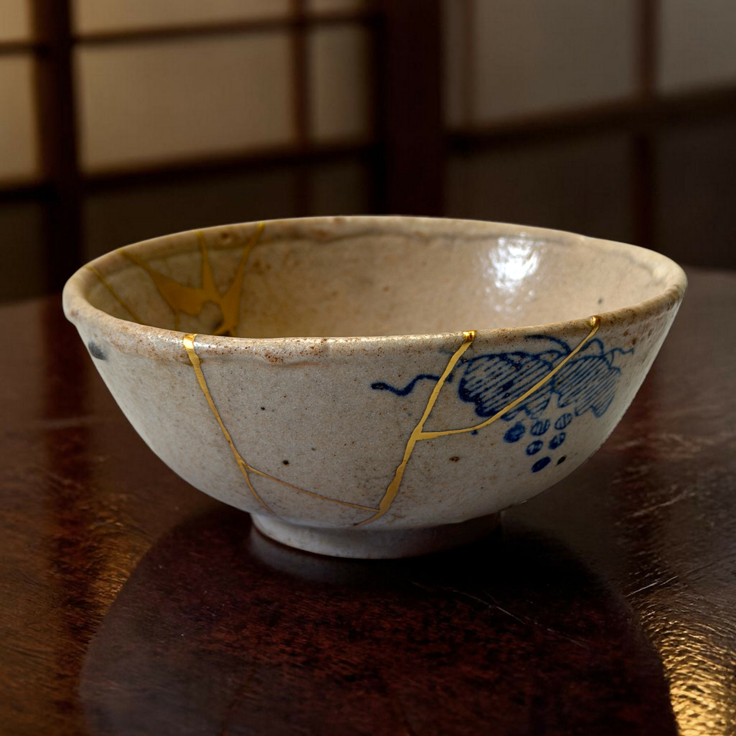 IMG_1579-Photoroom | Kintsugi by Myriam GREFF Close-up of a ceramic bowl with blue glaze designs and gold streaks on a dark wooden table.