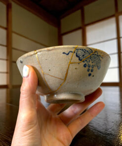 Close-up of a hand holding a speckled ceramic bowl with blue patterns and gold repair lines, indoors against wood paneling.