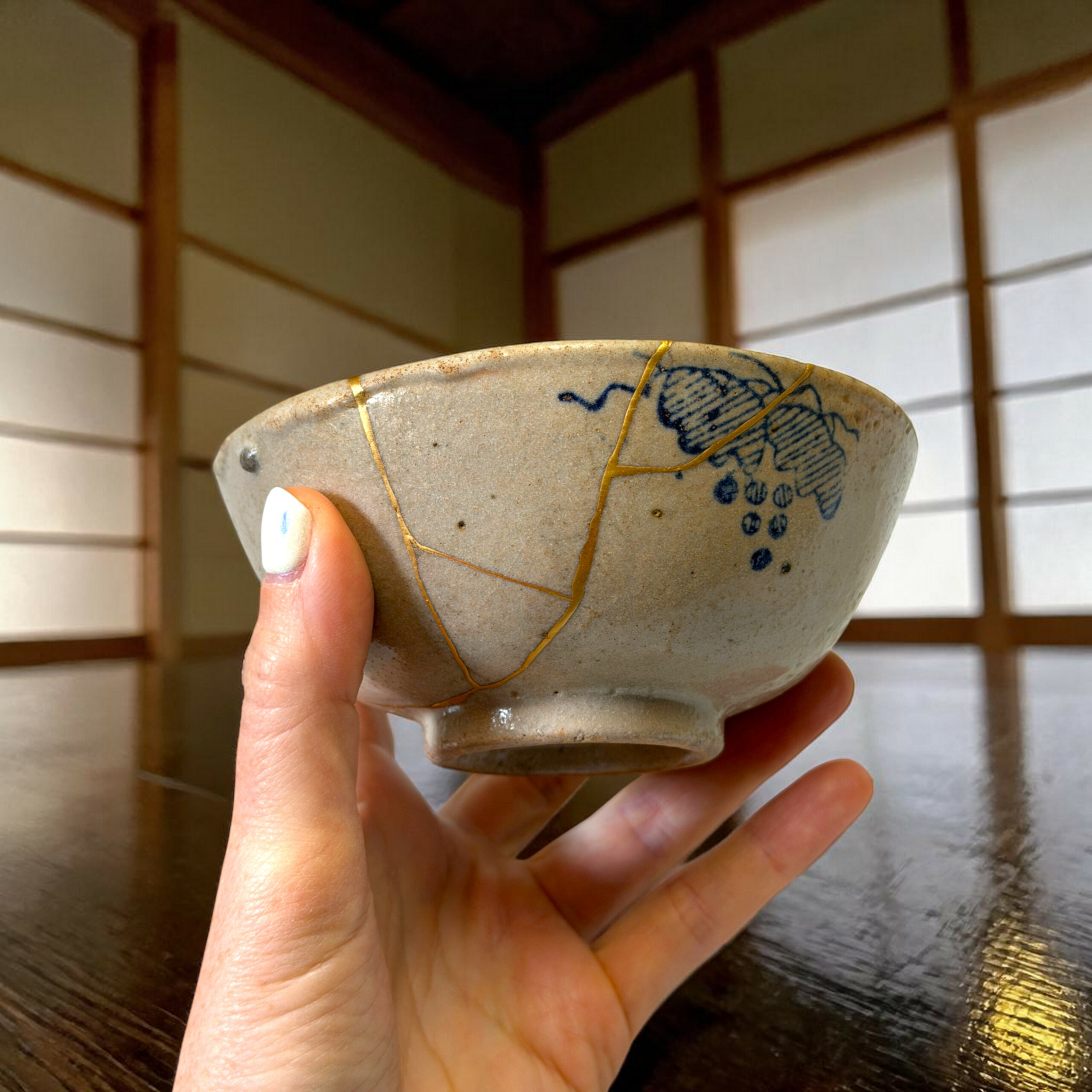 IMG_1580-Photoroom | Kintsugi by Myriam GREFF Close-up of a hand holding a speckled ceramic bowl with blue patterns and gold repair lines, indoors against wood paneling.