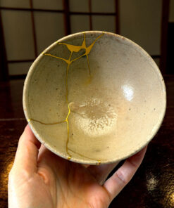 Close-up of a beige ceramic bowl repaired with gold lacquer (kintsugi) held in a person's hands over a dark wooden surface, highlighting the crack lines and texture.