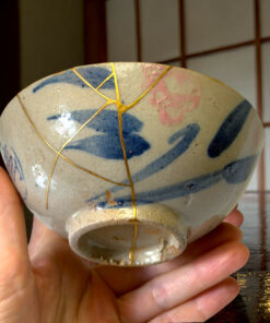 Close-up of a ceramic vase with gold kintsugi cracks, held in two hands against a dark background.