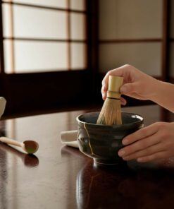 Hands whisking matcha in a dark ceramic bowl with a gold-kintsugi crack, on a polished wooden table.