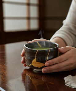 Hands cradle a steaming ceramic tea bowl with gold glaze on a polished wooden table