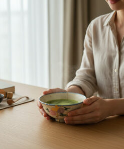Person in a light shirt holds a ceramic bowl with green tea at a wooden table, with a whisk and cup nearby in a calm setting.