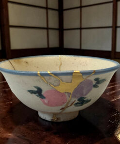 Close-up of a ceramic bowl with a blue rim, floral design, and visible gold kintsugi repairs on a dark wooden table, background lattice panel