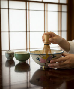 Person whisking matcha in a decorated ceramic bowl with a bamboo whisk on a dark wooden table.