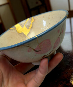 Close-up of small painted ceramic bowl held in a person's hands, featuring a blue rim and yellow decorative shapes.