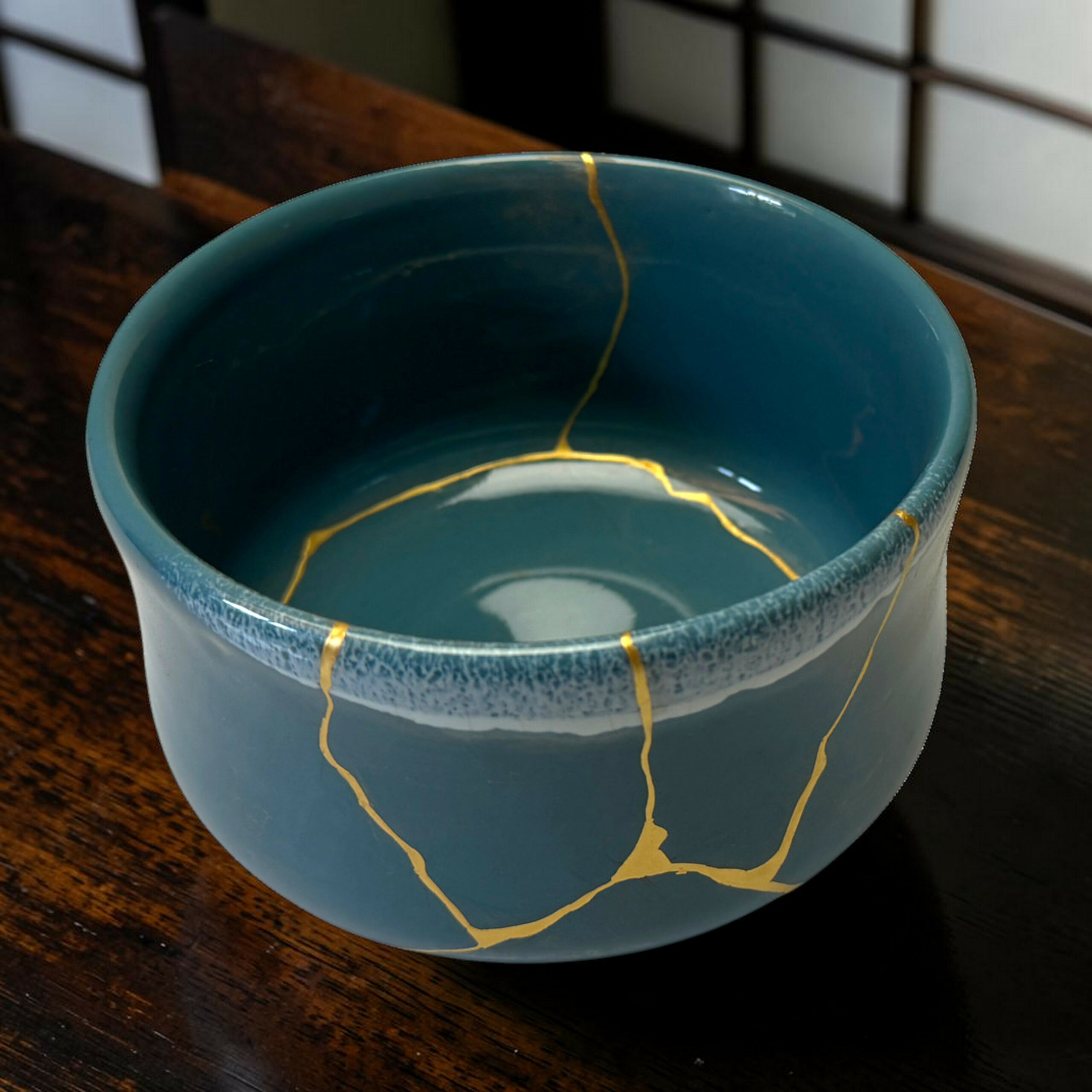 IMG_1676-Photoroom | Kintsugi by Myriam GREFF Close-up of a blue ceramic mug with gold glaze marks running along its rim, set on a wooden surface.