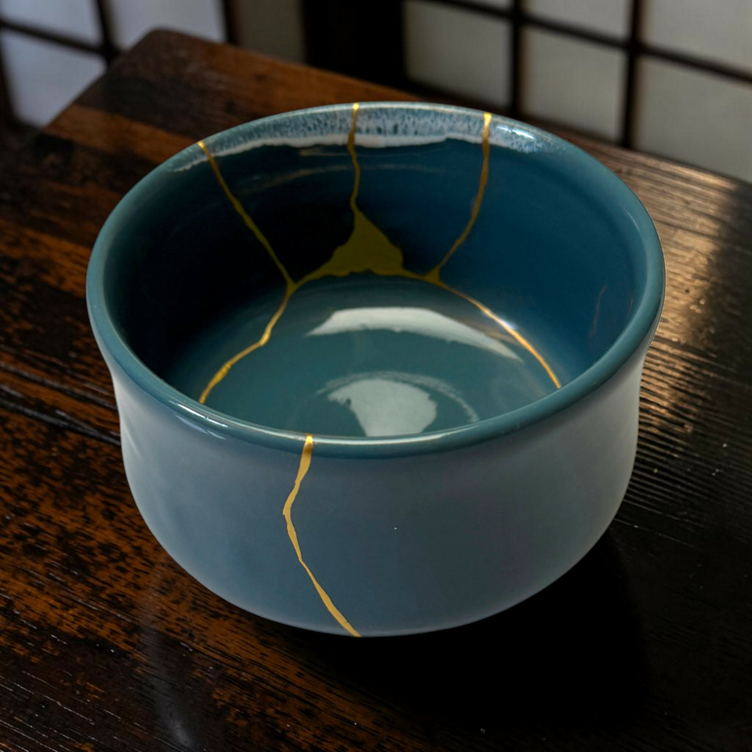 IMG_1678-Photoroom | Kintsugi by Myriam GREFF Close-up of a teal ceramic mug with yellow crackle glaze along the rim, placed on a dark wooden table.