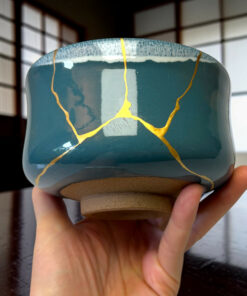Close-up of a teal ceramic mug with yellow crackle lines being held by a hand over a dark wooden table.