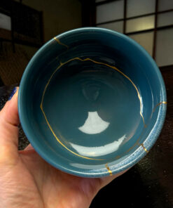 Close-up of a teal ceramic bowl being held by a hand, with gold streaks inside the bowl, on a dark wooden surface.