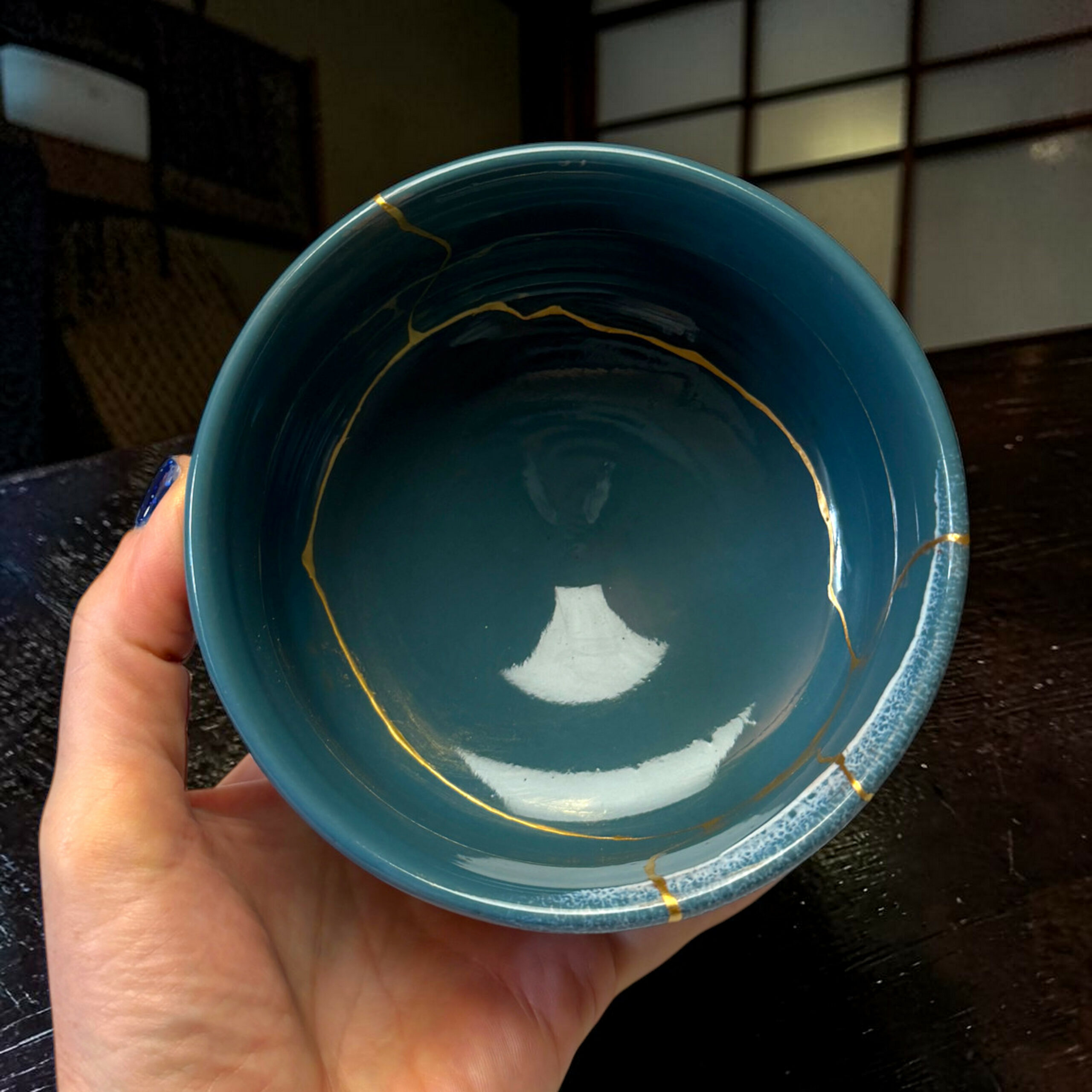 IMG_1680-Photoroom | Kintsugi by Myriam GREFF Close-up of a teal ceramic bowl being held by a hand, with gold streaks inside the bowl, on a dark wooden surface.