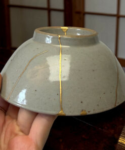 Close-up of a gray ceramic vase with gold kintsugi repairs along the cracks, held in a person’s hands.