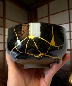 Close-up of a black ceramic pot with a gold crackle pattern being held by hands in a wooden-room background.
