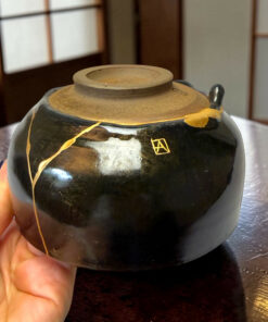 Close-up of a dark ceramic teapot with a dusty round lid, tied with yellow string, being held over a wooden table by a hand