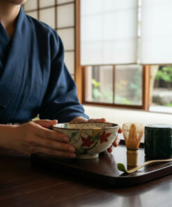Person in a dark blue kimono holds a decorated ceramic tea bowl on a tray, with a bamboo whisk and tea canister nearby by a window.
