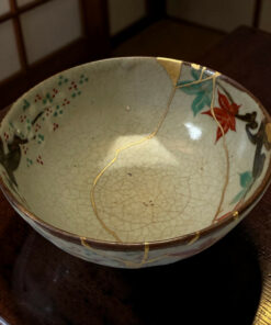 Close-up of a cracked ceramic bowl repaired with gold lacquer (kintsugi), showing a pale glaze with red and green floral patterns on a dark wooden table.