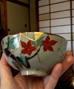 Decorative ceramic bowl with red flowers and green leaves held by hands, with a wooden lattice screen in the background.