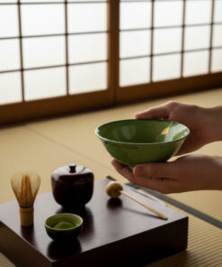 Person holds a green ceramic bowl during a Japanese tea ceremony setup on a dark wooden platform with a whisk, small bowl of matcha, and a teapot nearby on tatami mats in the background