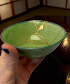 Close-up of a green ceramic bowl with gold crackle lines, held by a hand with blue-painted nails, on a wooden table background.