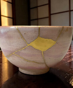 Close-up of a pale ceramic pot with gold kintsugi cracks resting on a dark wooden surface.
