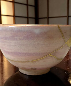 Close-up of a lavender ceramic bowl with a yellow string draped over its rim on a dark wooden table, background blurred with paneling.