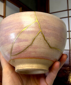 Close-up of a hand holding a beige ceramic bowl with gold cracks from a kintsugi repair, set against a wooden floor background.