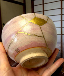 Close-up of a pale round object with gold-filled cracks (kintsugi) held in a person's hand over a wooden surface.