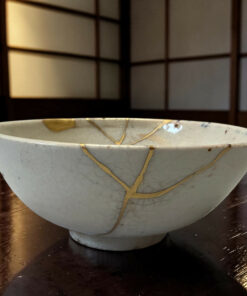 White ceramic bowl repaired with gold seams (kintsugi) on a dark wooden table. The edge of another bowl is faintly visible behind it.