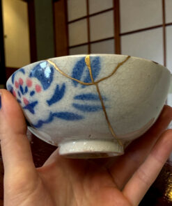 Close-up of a blue-and-white ceramic bowl with gold lines being held by a hand, Japanese-style shoji screen in the background.