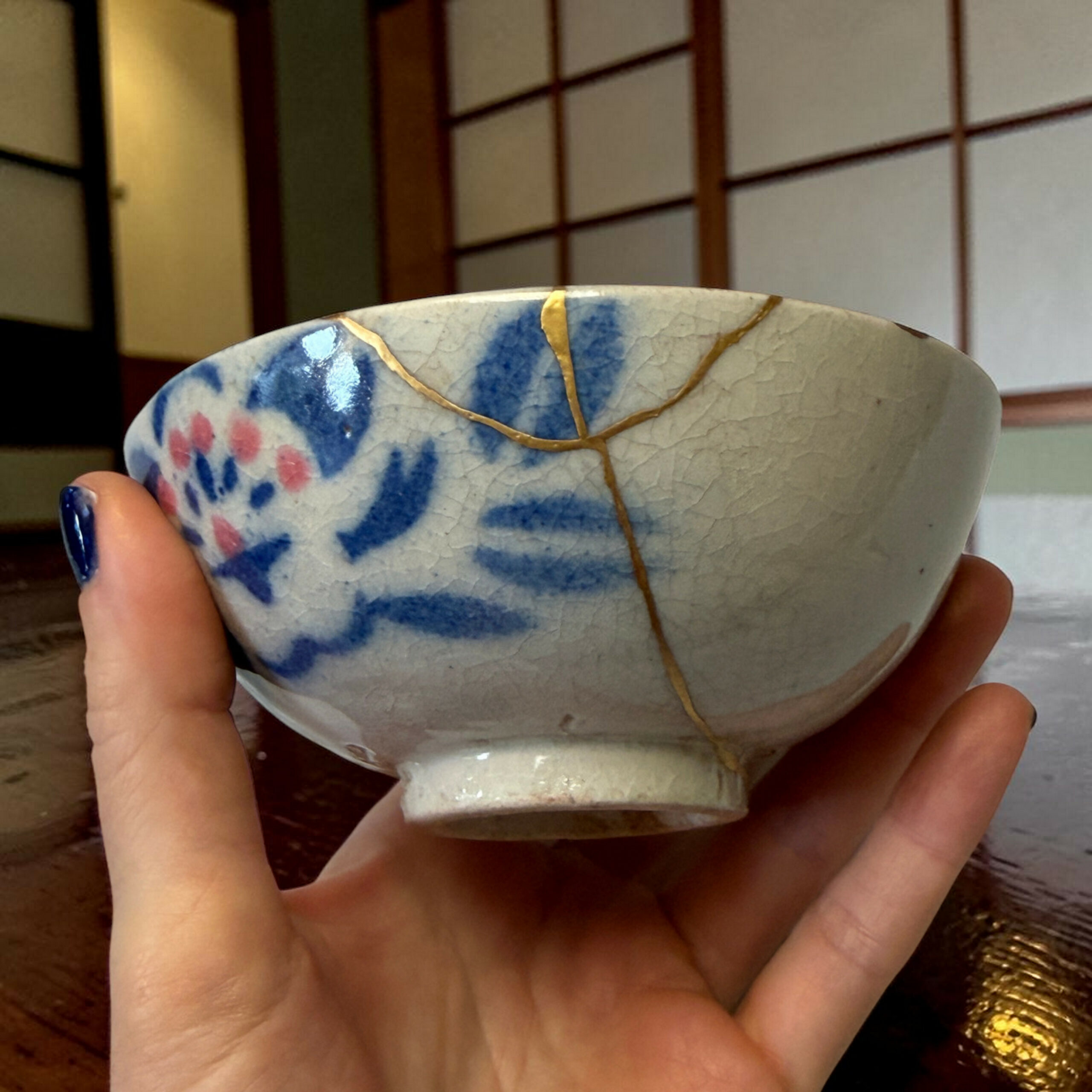 IMG_1715-Photoroom | Kintsugi by Myriam GREFF Close-up of a blue-and-white ceramic bowl with gold lines being held by a hand, Japanese-style shoji screen in the background.