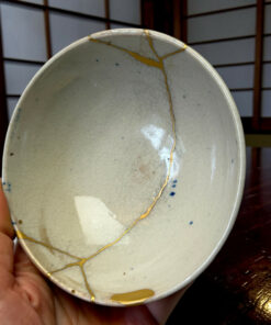 A ceramic plate with visible gold repair lines (kintsugi) being held by a hand, with a dark wooden table in the background.