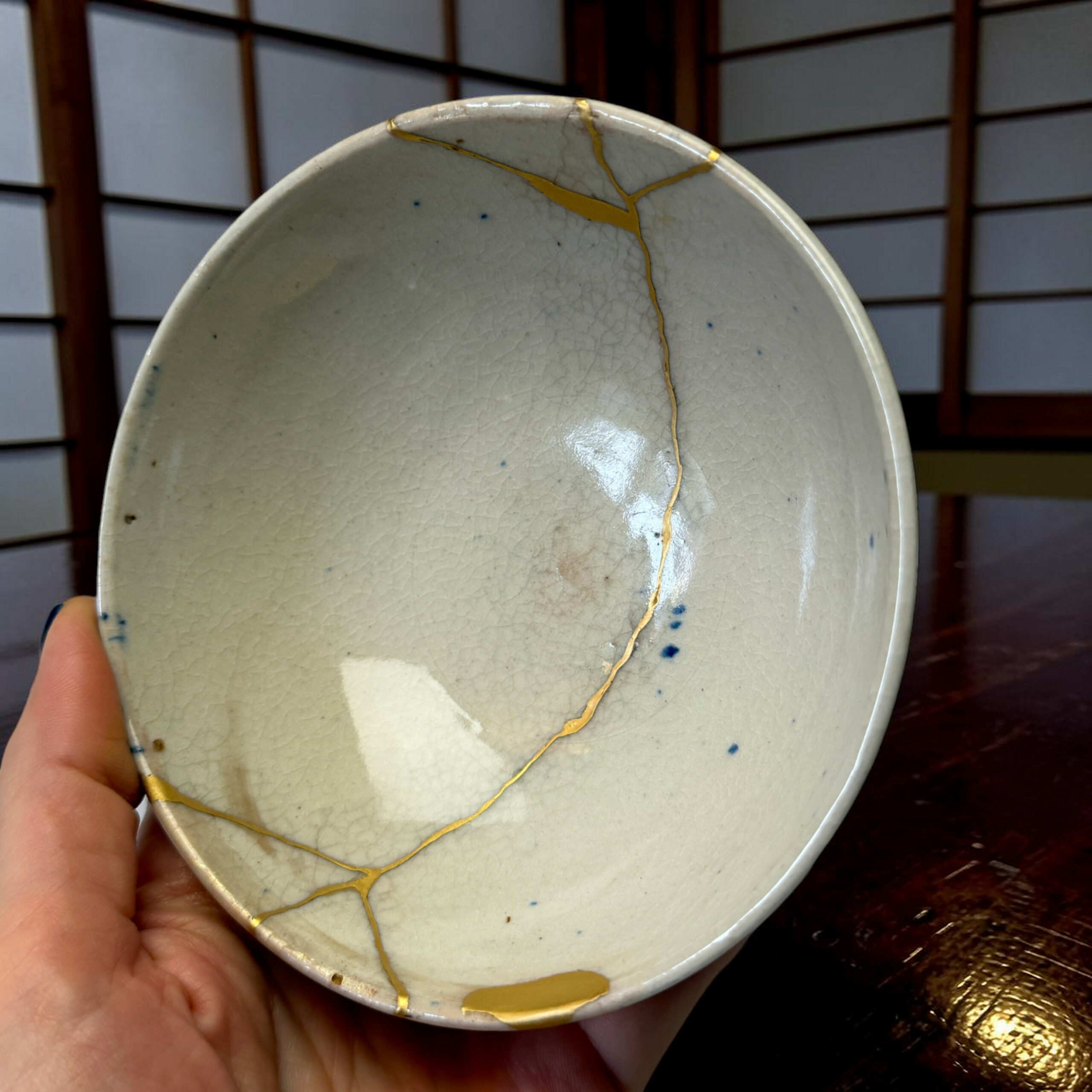 IMG_1716-Photoroom | Kintsugi by Myriam GREFF A ceramic plate with visible gold repair lines (kintsugi) being held by a hand, with a dark wooden table in the background.