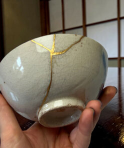Hands cradle a large smooth gray ceramic sphere with gold cracks (kintsugi) against a wooden surface.