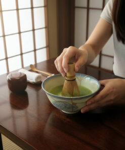 Person whisking vibrant green matcha powder in a ceramic bowl during a tea ceremony at a wooden table, natural light streaming through shoji screens
