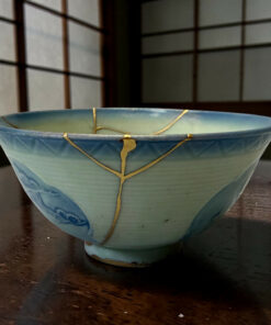 Close-up of a light blue ceramic bowl with gold strings tied around its rim on a dark wooden table.