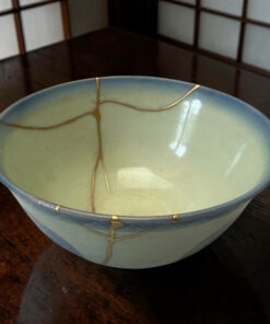 Close-up of a light blue ceramic bowl with gold kintsugi cracks on a dark wooden surface.