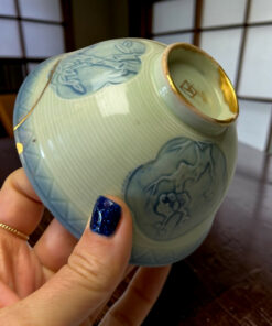 Close-up of a pale blue ceramic vase with floral relief and a gold rim, held by a hand with dark blue glittery nails.