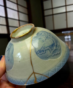 Close-up of a light blue ceramic vase with blue floral patterns and gold trim, held by a finger against a blurred background shelf.