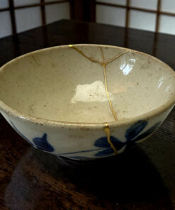 Close-up of a cream ceramic bowl with gold repair seams on a dark wooden table; a blurred shoji-style screen in the background.