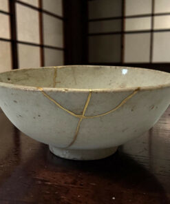 A light gray ceramic bowl with a pale string wrapped around its rim sits on a dark wooden table, background blurred geometric panels.