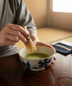 Person whisking matcha in a blue-and-white ceramic bowl using a bamboo chasen on a wooden table.